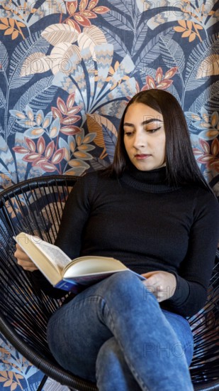 A young woman is engrossed in reading a book while sitting in a modern wicker chair, against a vibrant floral wallpaper background