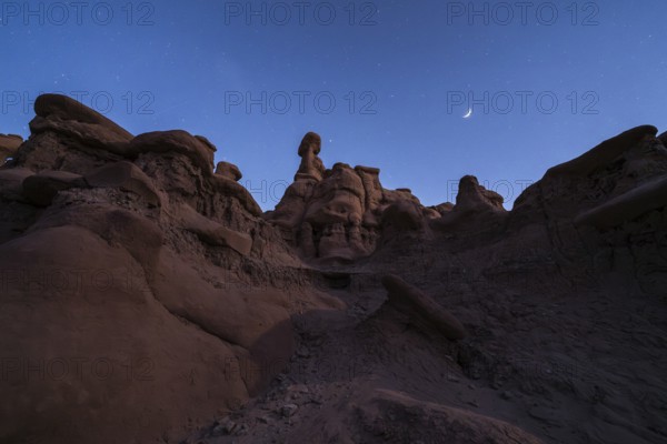 A serene night view of sandstone formations under a starry sky with a crescent moon in Goblin Valley State Park, Utah. The natural beauty is otherworldly and majestic