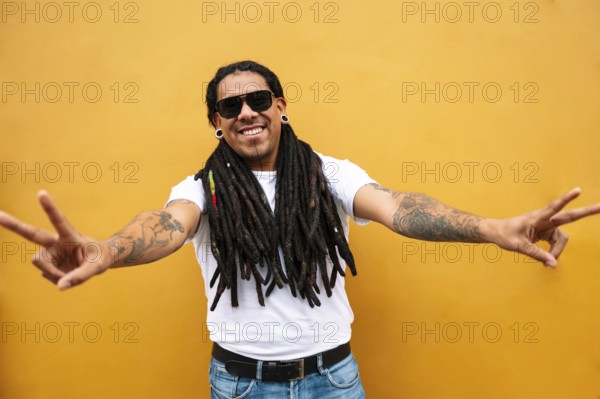 A cheerful man with long dreadlocks and tattoos poses against a vibrant yellow wall in Barranco, Lima, Peru, making a peace sign with both hands