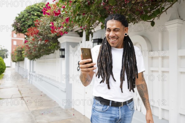 A joyful man with dreadlocks holds a smartphone, smiling widely as he stands in front of a vibrant, floral-adorned wall in Barranco, Lima, Peru, capturing a moment of happiness