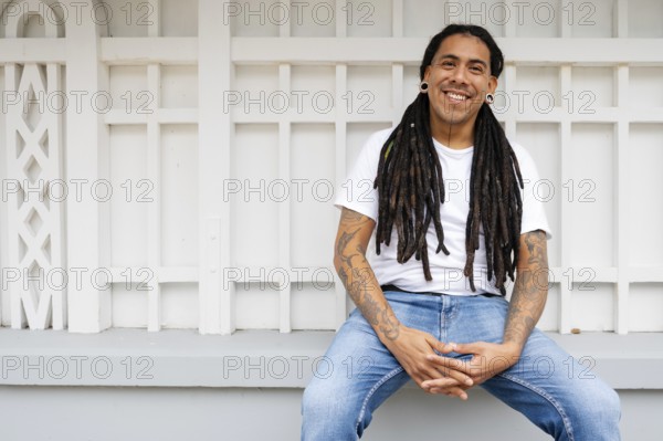 A cheerful man with long dreadlocks and tattoos sits in Barranco, Lima, Peru, exuding a relaxed vibe He wears a white shirt and jeans, enjoying a sunny day outdoors
