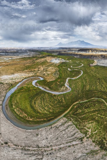 A breathtaking aerial drone photograph captures a winding river snaking through the rugged terrain of Hanksville, Utah, showcasing the contrasting verdant riverbanks and arid landscape under a dynamic sky
