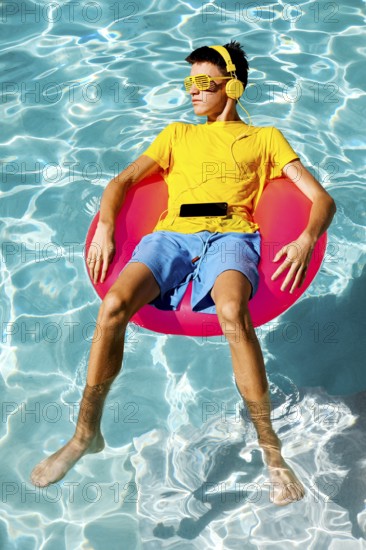 A young teenage boy in vibrant summer attire floats on a pink inflatable ring in clear blue pool water, wearing yellow headphones and shutter shades, enjoying music in the sun