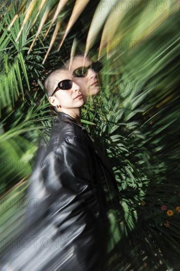 A stylish lesbian woman wearing sunglasses and a leather jacket stands amidst lush green foliage. The image features a unique blurred motion effect, enhancing the dynamic composition