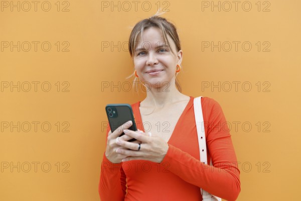 A woman stands against an orange wall in Warsaw, intently using her smartphone Dressed in a red top with matching earrings and a white bag, she appears engaged and focused