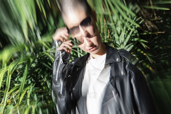 A stylish lesbian woman in a leather jacket and sunglasses is partially blurred against a backdrop of lush green foliage, capturing a vibrant, dynamic outdoor scene
