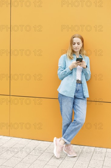 A woman leans against a bright orange wall in Warsaw, focused on her phone She wears a blue shirt, jeans, and sneakers The scene portrays urban style and digital life