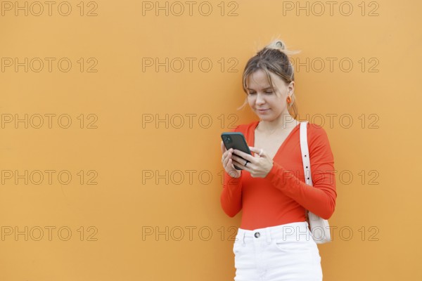 A woman in a vibrant orange outfit uses her smartphone against a matching orange background in Warsaw Her focus and style reflect modern urban life and technology