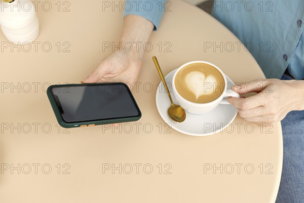 A woman relaxes in a Warsaw cafe, holding a smartphone and a cup of coffee with latte art The cozy setting and modern gadgets create a peaceful atmosphere