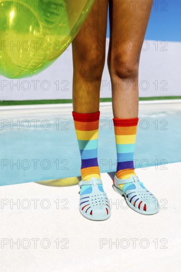 Vibrant scene featuring legs in rainbow socks and teal sandals, beside a pool. The bright turquoise water and inflatable float enhance the summer feel and colorful theme