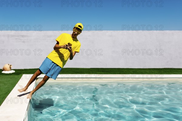 A teenage boy in vibrant yellow attire leans playfully by a pool, capturing a moment with a phone. The clear blue sky and pristine pool create a lively summer vibe