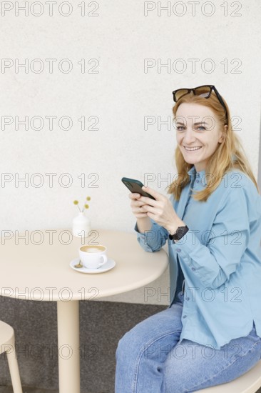 A smiling woman enjoys coffee while using her smartphone at a cafe in Warsaw She wears casual clothing and sunglasses, seated at a cozy outdoor table