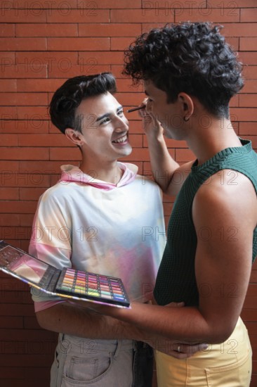 A couple shares a tender moment as one applies makeup to the other with a colorful palette, set against a brick wall, expressing love and creativity