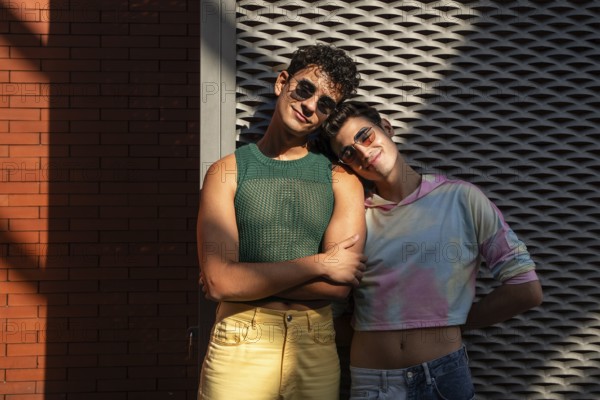A joyful gay couple in stylish outfits stands together, one leaning on the other affectionately. They're enjoying a sunny day, with a red brick wall as a backdrop