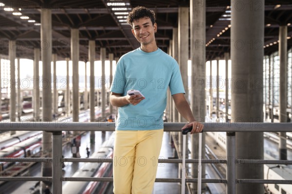 A young traveler in a vibrant blue shirt leans casually against a railing at a train station, checking his phone. Luggage and trains are visible beneath the high ceiling