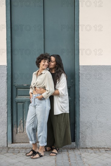 A loving lesbian couple stands in front of a teal door, embracing warmly. One partner gently kisses the other's cheek, capturing a moment of love and affection