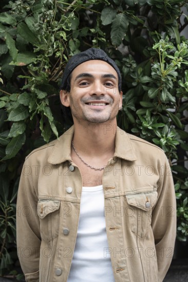 A gay man in casual clothing stands against a lush, leafy background. He sports a chain necklace and earring, conveying a relaxed and stylish vibe in natural surroundings