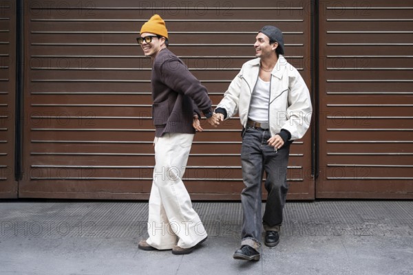 A joyful couple holds hands while walking in an urban setting. They are smiling, showcasing happiness and connection. One wears a beanie, the other a cap, exuding casual style
