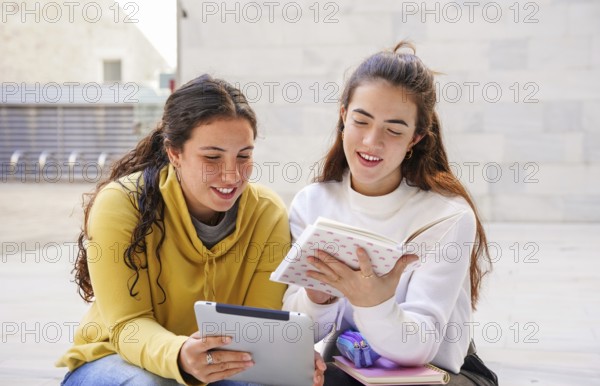 Two students sit together outdoors, enjoying a collaborative study session One holds a tablet, while the other reads a book, fostering learning and connection
