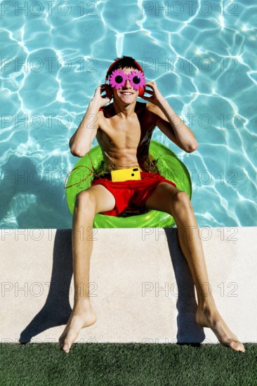 Young teenage boy sits on a green float in a pool, wearing vibrant red shorts, flower-shaped pink sunglasses, and headphones, embodying the essence of summer fun and relaxation