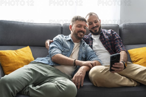 A joyful gay couple sits comfortably on the sofa, smiling as they look at a smartphone together in a cozy living room