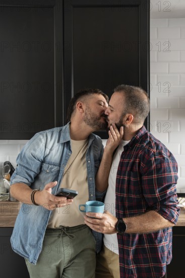 A loving gay couple shares a tender kiss in their kitchen, creating a warm and intimate atmosphere. One holds a cup, the other a phone, dressed in casual shirts