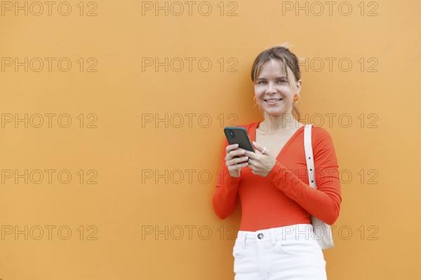 A woman wearing a red top stands against an orange wall, holding a smartphone The vibrant colors and urban setting capture a modern lifestyle in Warsaw