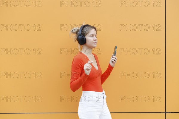 A young woman wearing headphones, joyfully listening to music and holding a smartphone She stands against a vibrant, orange wall in Warsaw, exuding a carefree vibe