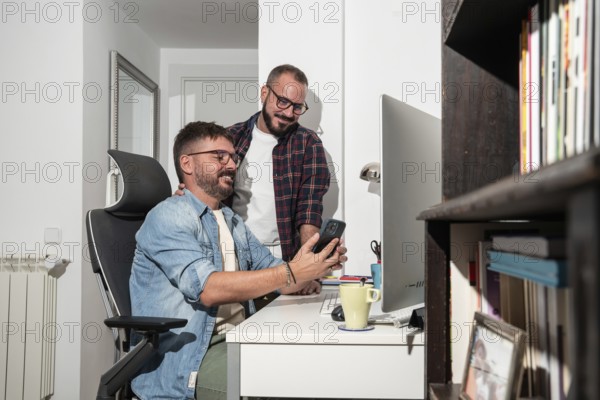 A joyful gay couple shares a moment at their home office. One man sits at a desk, holding a phone, while the other stands beside, smiling warmly, with books in the background