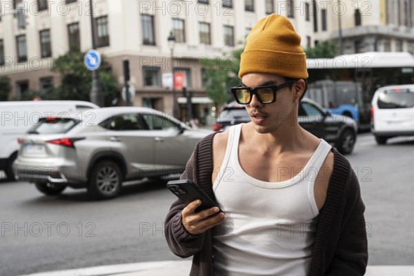 A Colombian man stands on a busy city street, wearing stylish sunglasses and a beanie as he checks his smartphone amidst bustling traffic and buildings