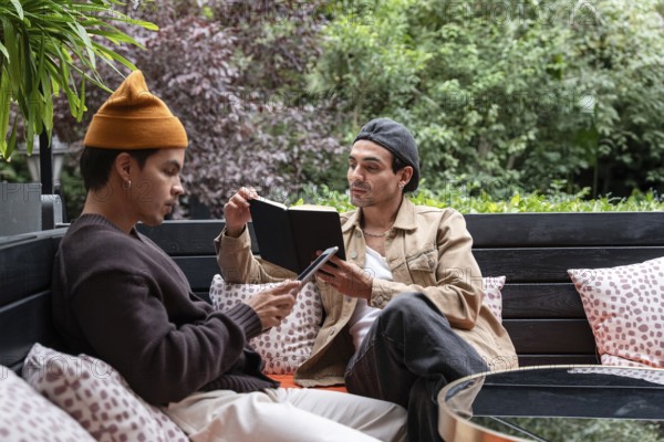 A Colombian gay couple enjoys a quiet day outdoors on a cozy bench. One reads from a book, while the other engages with a tablet, surrounded by lush greenery