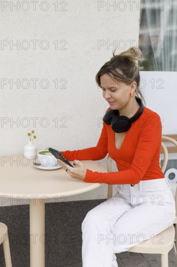 A woman sits at an outdoor cafe in Warsaw, using her smartphone and wearing headphones She enjoys a cup of matcha while engaging with her gadgets in a relaxed environment