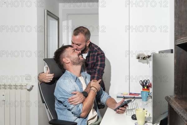 A joyful gay couple shares an affectionate moment in their home office. One partner sits with a phone while the other embraces him from behind, creating a warm atmosphere