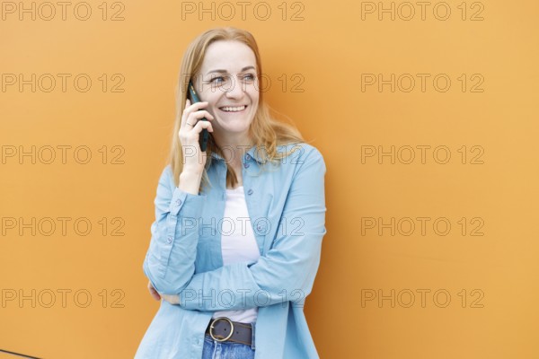 Smiling woman stands against a vibrant orange wall in Warsaw, chatting on a smartphone She wears casual blue and white attire, embodying a relaxed, modern vibe