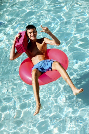 Young teenage boy enjoys a sunny day in a swimming pool, lounging on a pink inflatable ring. He holds a radio, smiling and gesturing a peace sign, capturing the essence of summer leisure