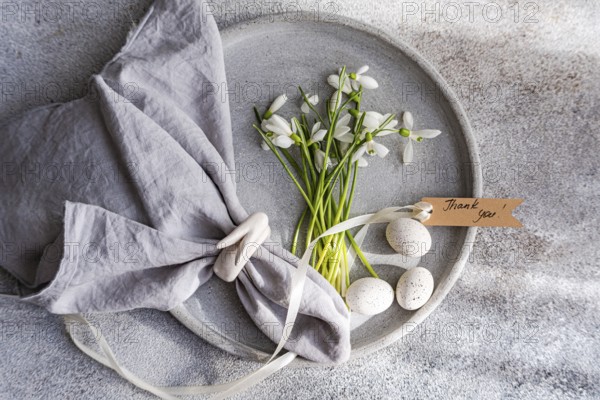 Top view of an exquisite spring table setting featuring fresh snowdrops arranged on a gray ceramic plate, complemented by a gray linen napkin, delicate quail eggs, and a Thank you! tag, all laid on a textured surface
