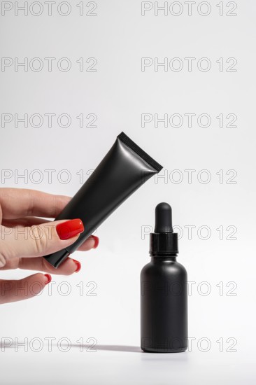 Cropped unrecognizable female hand with red manicure holding a matte black tube and dropper bottle isolated on a white background