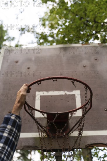 A hand reaches up to grab an old, iron basketball hoop with a chain net in an outdoor area surrounded by greenery in Sweden. The rustic charm highlights simplicity and nostalgia