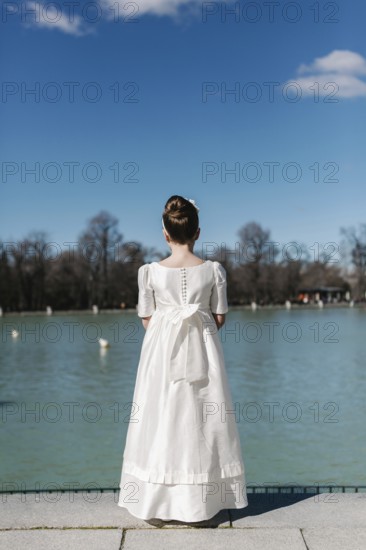 A girl stands by a serene lakeside, wearing a beautiful white First Communion Day dress. Her hair is elegantly styled, capturing simplicity and grace