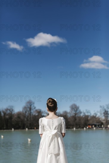 A young girl in a white First Communion dress stands by a tranquil lake, gazing at the serene view. The clear blue sky and peaceful water create a sense of calm