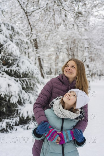 A joyful mother embraces her daughter during a snowy winter day. They both wear colorful winter clothes, surrounded by snow-covered trees, creating a serene and cheerful scene