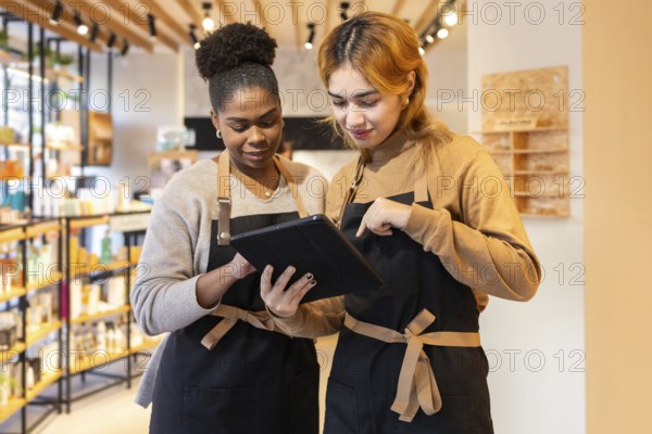 Two employees in a cosmetics store wearing aprons collaborate on a digital tablet, discussing product details. Shelves filled with diverse beauty products adorn the background