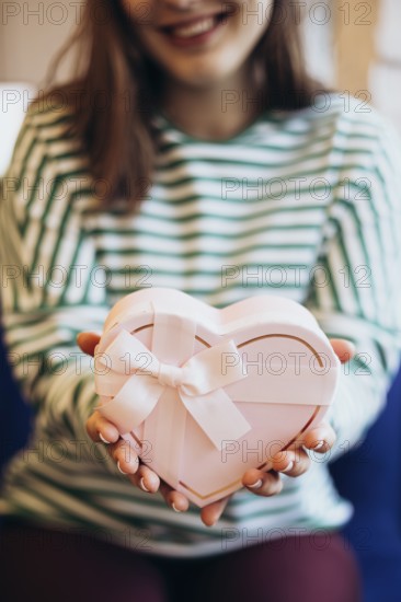 Cropped unrecognizable woman in a striped top holding a heart-shaped gift box adorned with a white ribbon, symbolizing love and generosity