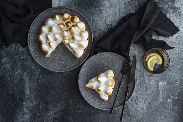 A delectable homemade lemon cake with toasted meringue, served on gray plates. Black cutlery and a glass with lemon slices complete the elegant table setting on a textured surface