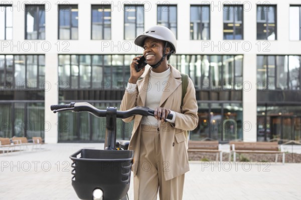 Smiling black woman in sleek beige suit and helmet, talking on a smartphone while holding a bike in a modern urban environment Confident and stylish city life