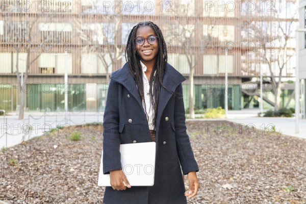 Smiling African American businesswoman, dressed in a stylish coat and glasses, holds a laptop while enjoying a break outdoors with modern buildings in the background