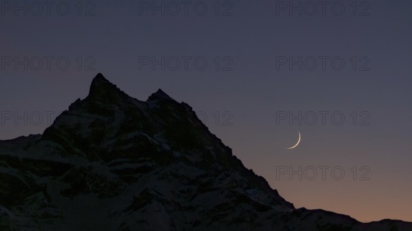 The crescent Moon gracefully hangs over the silhouette of the Dents du Midi mountain in the Swiss Alps, set against a tranquil twilight sky, evoking both beauty and serenity