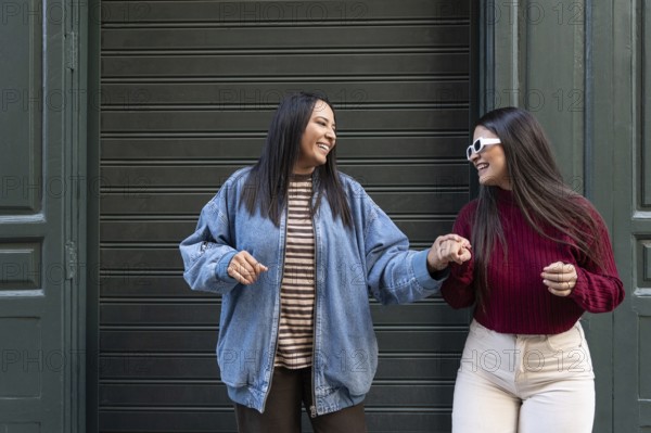 Joyful moment of a latin lesbian couple holding hands and laughing outside. Both wear casual clothes, displaying a relaxed, happy atmosphere against a neutral background