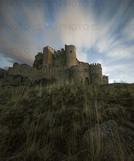 Majestic night view of Manqueospese Castle in Avila, Spain, with starry sky backdrop and wispy clouds
