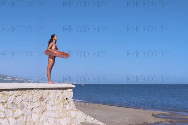 Side view of a young woman inside a float while standing atop a stone wall at the beach, gazing over a calm sea against a clear blue sky